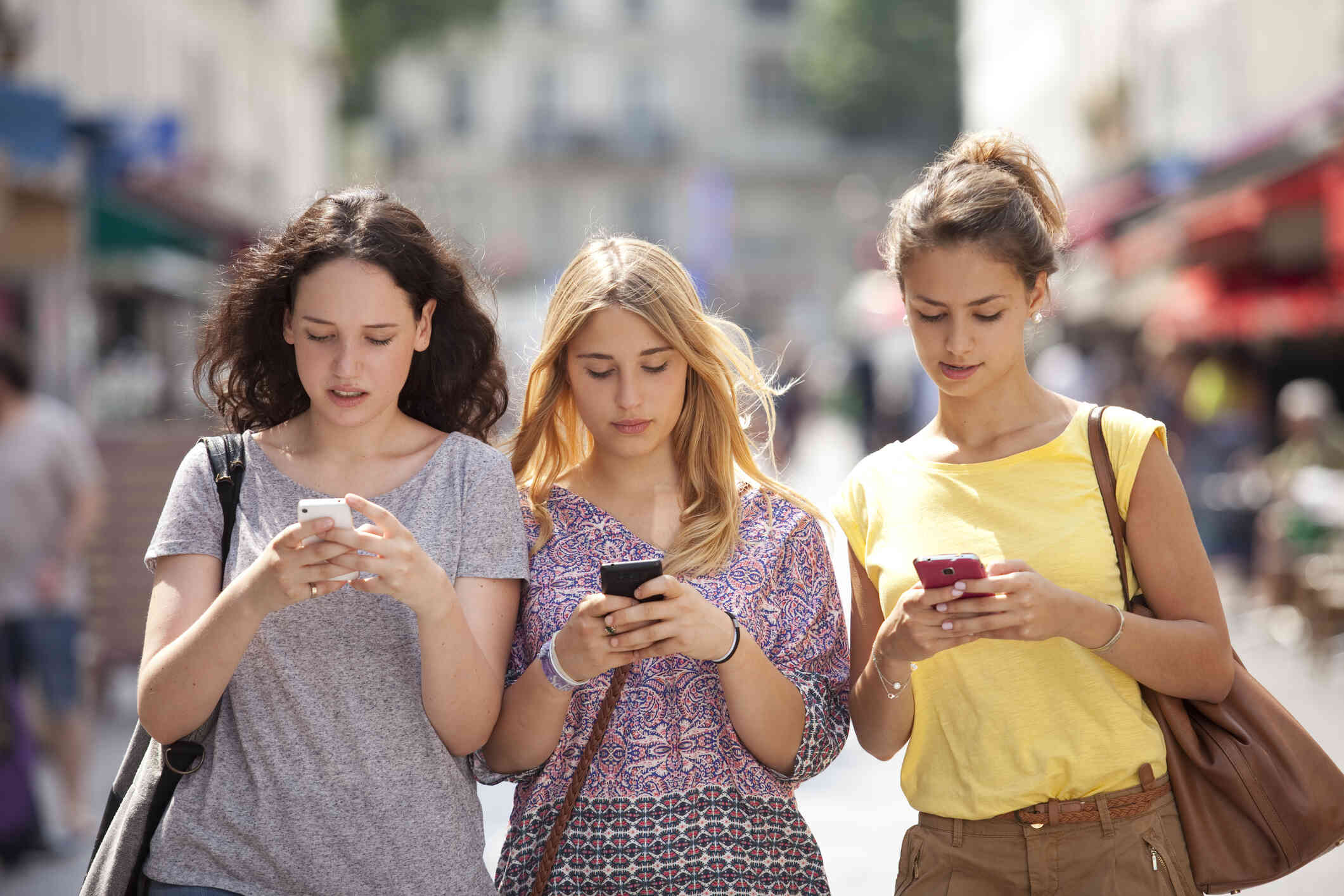 Three teenage girls walk down the street on a sunny day while looking down at the cellphones in their hands.